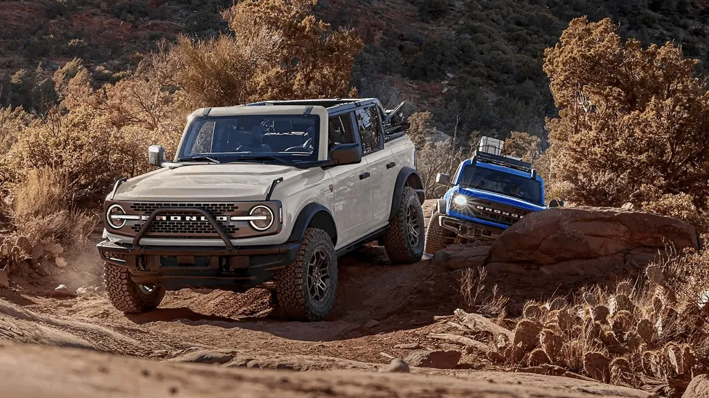 Ford Bronco 2026 gris clair sur un sentier rocailleux, suivi d'un second Bronco bleu.